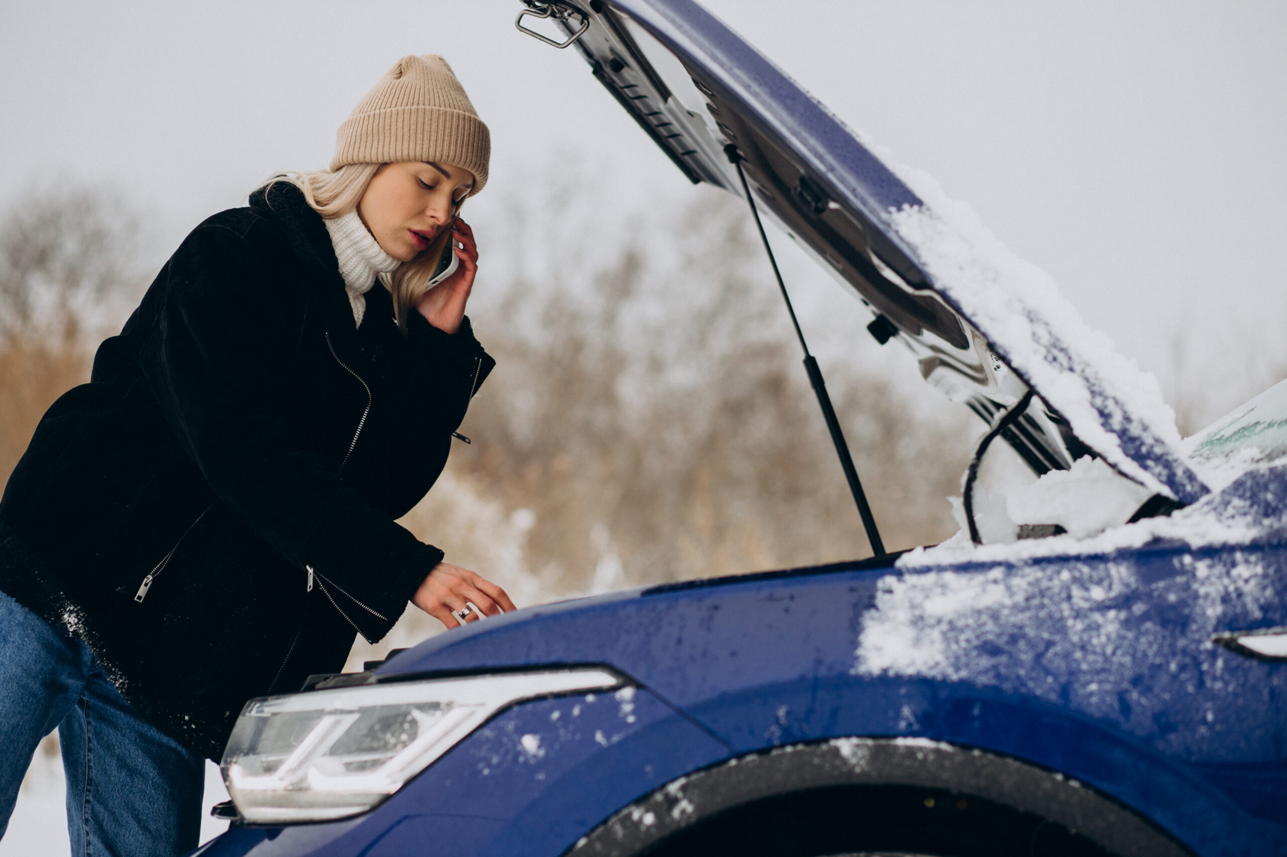 Mulher agasalhada falando ao celular ao lado do capô aberto de um carro azul na neve